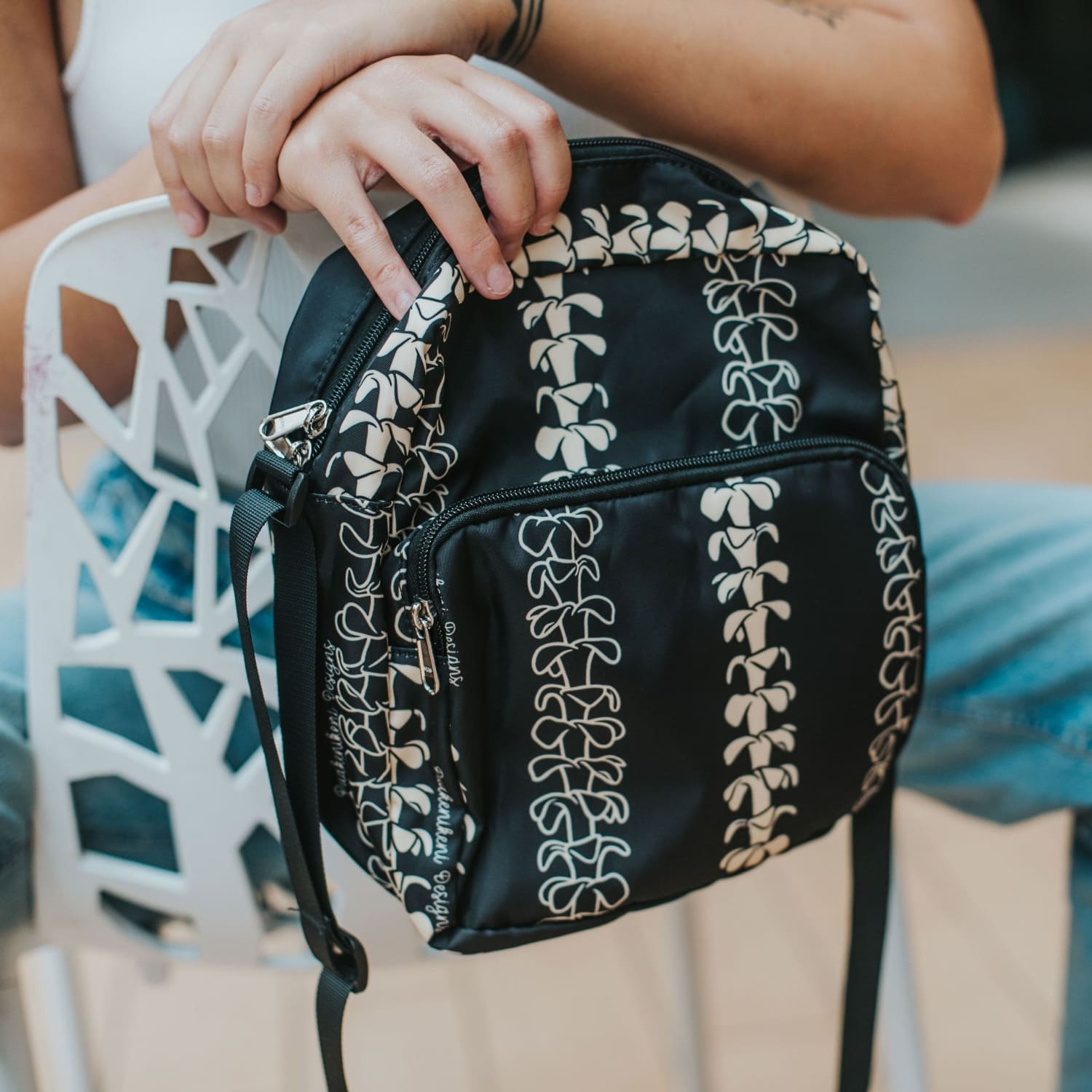 Crossbody bag by Puakenikeni Designs, in black with the best-selling pua kenikeni lei design, Kaulua; for women, moms, teenage girls, for travel and school Beige Lei Design model holding over chair close up